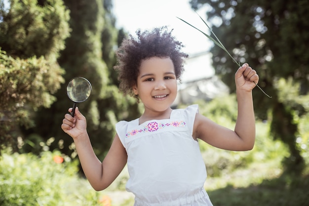 Good mood. Curly-haired little girl holding a magnifier and looking cheerful