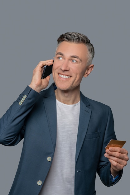 Good mood. Happy mature man with credit card talking on smartphone looking joyfully up and to side in studio photo