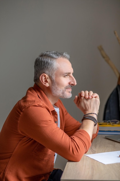 Good mood. A man in orange shirt sitting at the table and looking contented