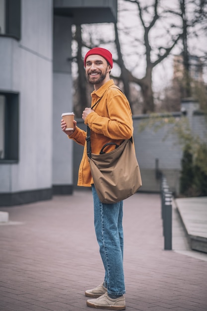 Good mood. Man in a red hat and orange jacket with a coffee cup in hands looking positive