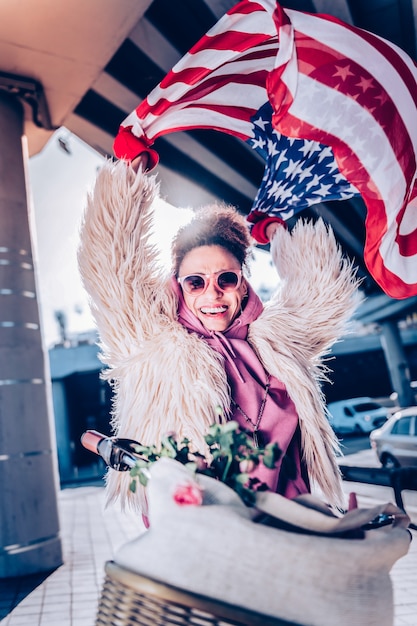 Good mood. Pleased international girl keeping smile on her face while demonstrating American flag