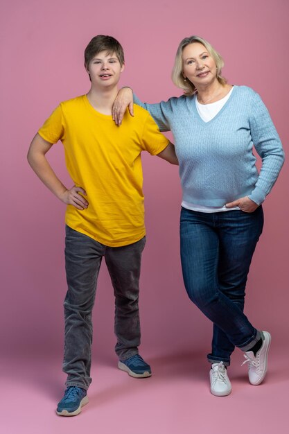 Photo good mood. satisfied guy with down syndrome holding hand on waist hugging smiling mom posing at camera on light background