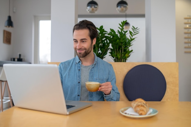 Хорошее настроение. Улыбающийся молодой бородатый мужчина с кофе сидит за столом с ноутбуком и смотрит на экран в кафе