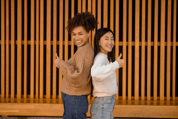 Good mood. Two smiling girls standing with their backs to each other looking at camera holding thumb up showing ok sign