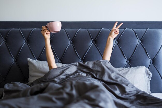 Good morning concept female hands with coffee mug and victory sign sticking out from the blanket