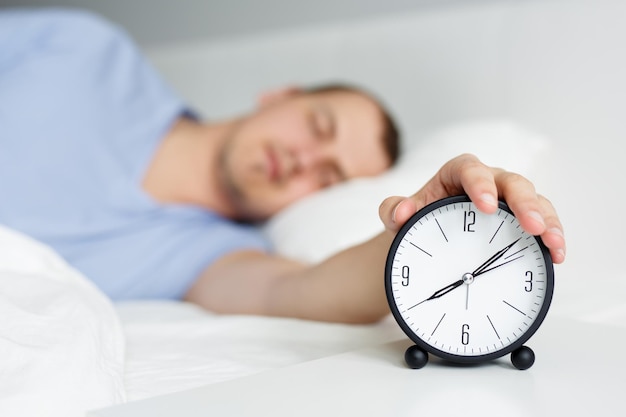 Good morning concept sleepy man turning off alarm clock on bedside table