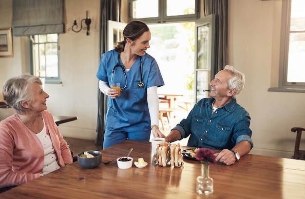 Доброе утро, мои любимые постояльцы. Снимок: молодая медсестра осматривает пожилую пару во время завтрака в доме престарелых.