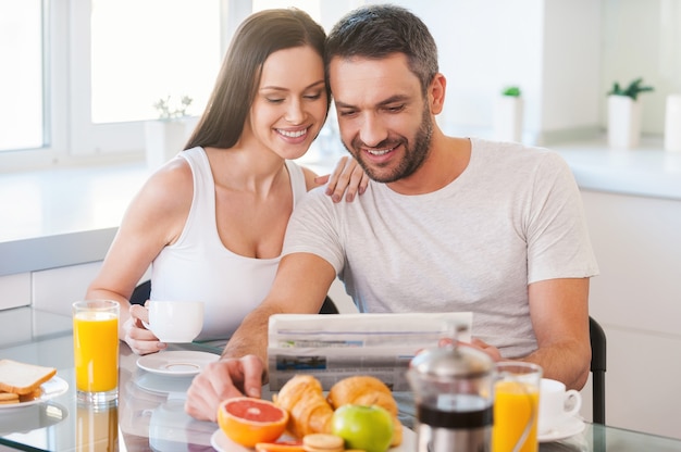 Good morning starts from good news. Beautiful young couple bonding to each other and reading newspaper together while sitting in the kitchen and having breakfast