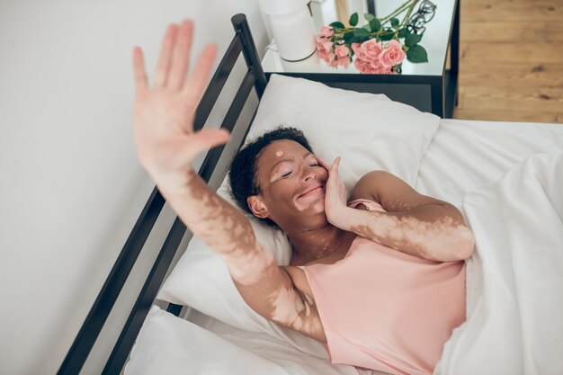 Good morning. A young dark-skinned woman lying in bed, stretching hand and smiling