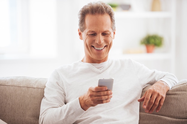 Good news from friend. Confident mature man holding mobile phone and looking at it with smile while sitting on the couch at home