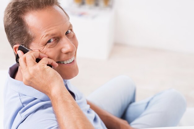 Good news from friends. Top view of cheerful mature man talking on the mobile phone and looking over shoulder while sitting on the couch at home