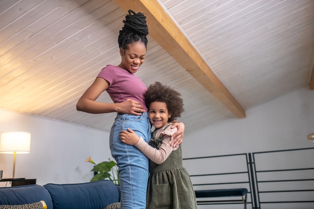 Good news. Little joyful african american girl touching her head to mothers belly standing hugging in room