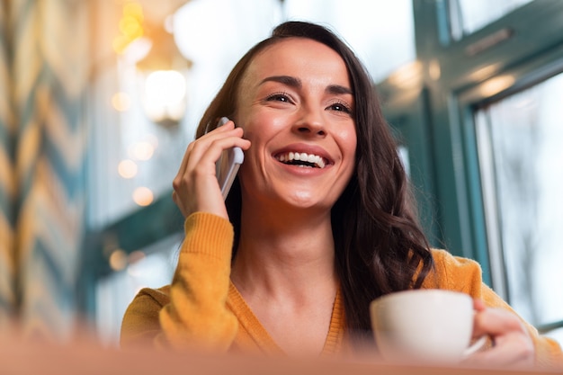 Good news. Low angle of jolly cute young woman calling boyfriend while posing on the blurred background and laughing