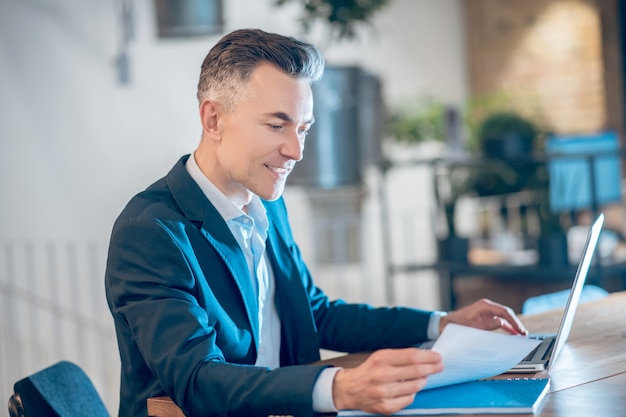 Good news. Smiling young adult business man in dark jacket sitting at laptop in office looking at paper document