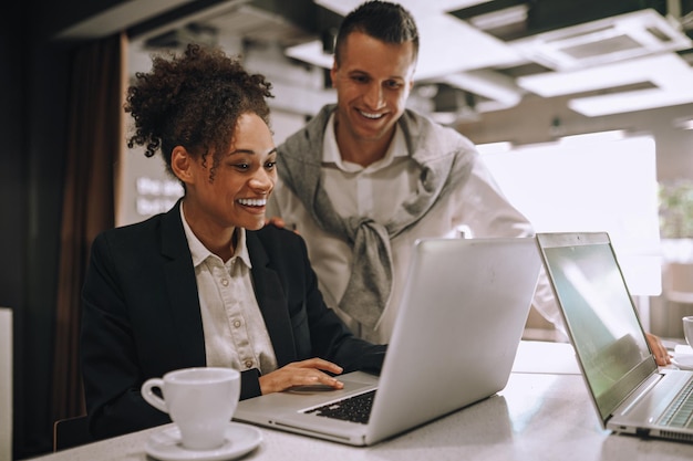 Photo good news smiling young adult mulatto woman sitting and caucasian man standing happily looking into laptop at workplace in office