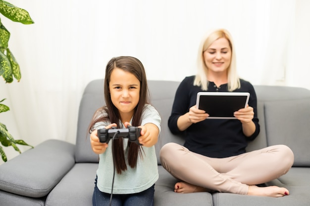 Good relationship cute little girl with young mother using\
joystick playing video game sitting together in living room\
enjoying family holiday.