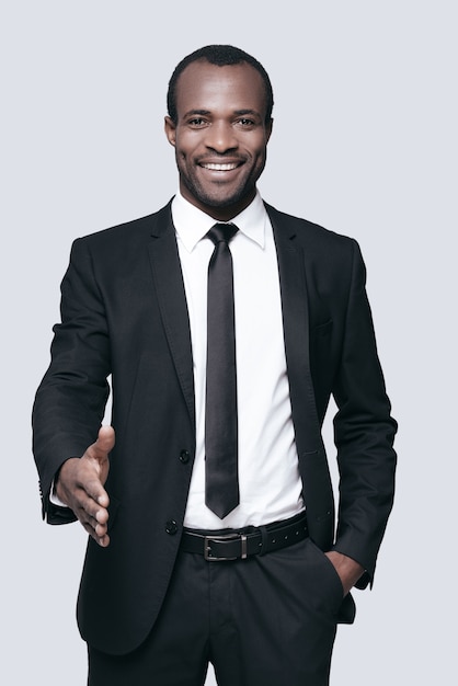 Good to see you in our team! Handsome young African man in formalwear man stretching out hand for shaking and smiling while standing against grey background