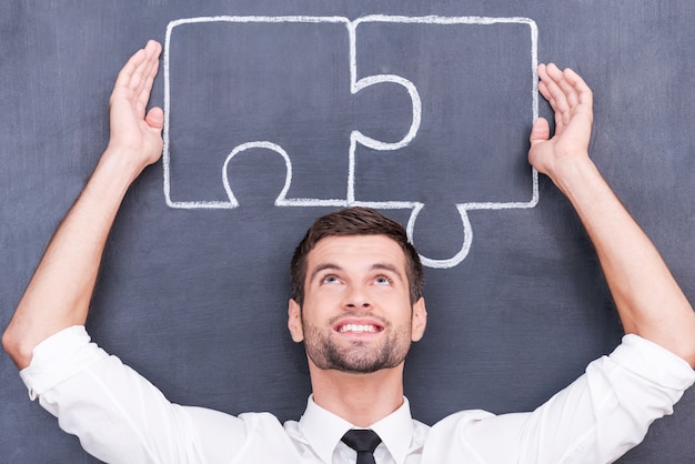 Good strategy - good solution. Handsome young man keeping arms raised and connecting jigsaw pieces on blackboard