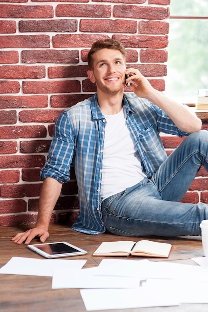 Good talk. Handsome young man sitting on the floor and talking on the mobile phone while digital tablet and documents laying near him