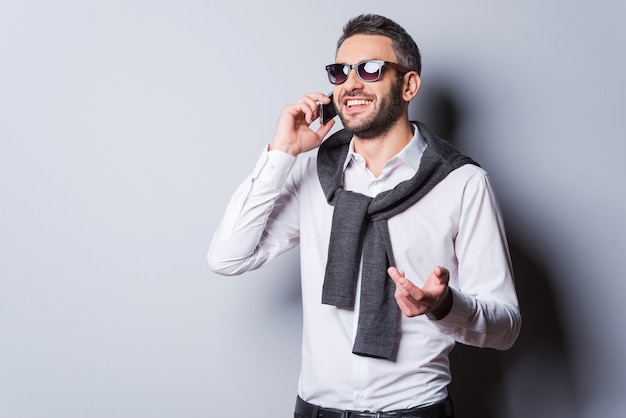 Good talk with friend. Handsome young man in smart casual wear and sunglasses talking on the mobile phone and smiling while standing against grey background