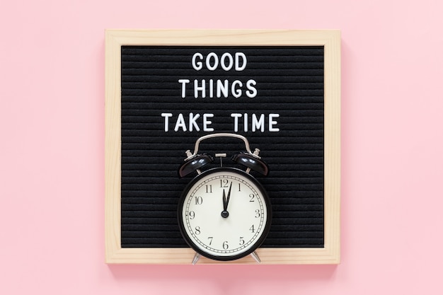 Good things take time. Motivational quote on black letter board, black alarm clock on pink background. Concept inspirational quote