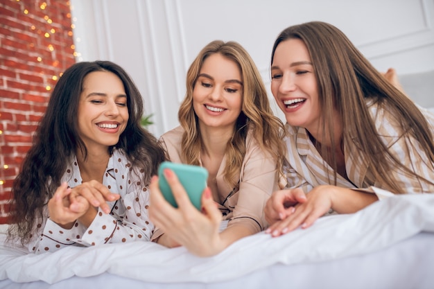 Good time. Three girls lying on bed and watching something on a smartphone