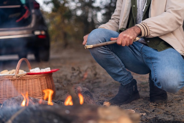 Good travel. Cropped view of the caucasian man spending time and roasting marshmallow around the forest. Their car at the background