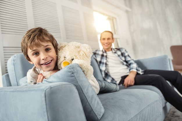 Good weekend. Attractive cheerful young fair-haired boy smiling and sitting on the couch with his toy and his daddy relaxing in the background