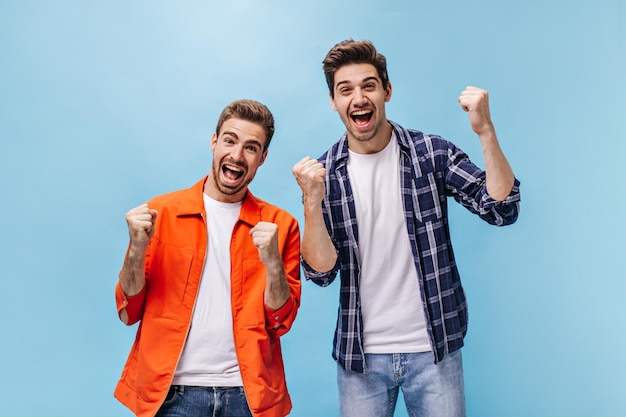 Goodhumored emotional men rejoice and celebrate victory on blue background Charming guy in orange jacket and brunet man in checkered shirt pose in great mood