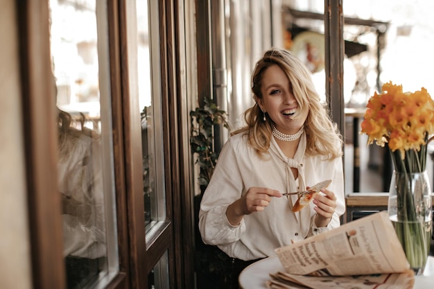 La giovane donna bionda di buon umore in camicetta bianca ed elegante collana di perle ride tiene coltello e pezzo di pane e si siede al ristorante