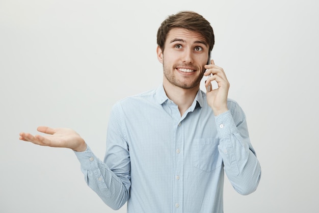 Bello giovane datore di lavoro felice in camicia casual che parla sullo smartphone facendo un gesto di pesatura con il palmo alzato che guarda in alto e sorride mentre esita