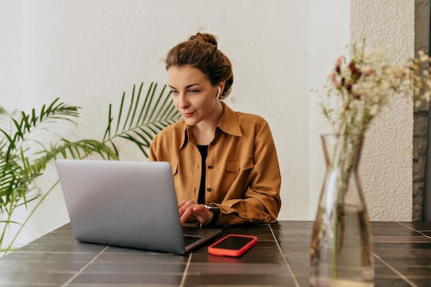 Bella giovane donna elegante con capelli raccolti e trucco nudo che indossa una maglietta digitando sul laptop nella caffetteria concetto di utilizzo e funzionamento a distanza