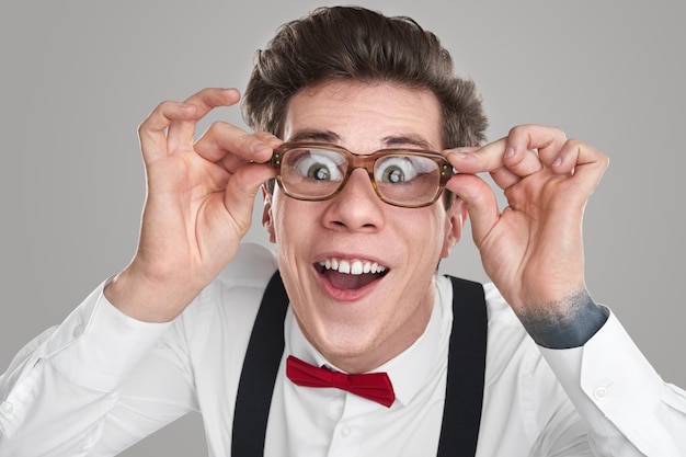 Goofy young man with huge eyes adjusting magnifying glasses and looking at camera with silly smile against gray background