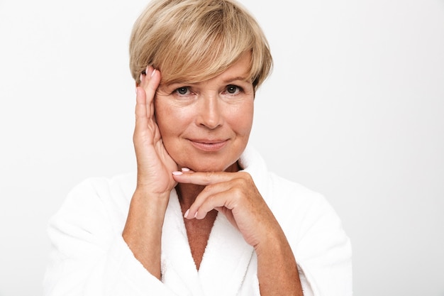 Foto splendida donna adulta con corti capelli biondi che indossa una vestaglia bianca che si tocca il viso isolato su un muro bianco