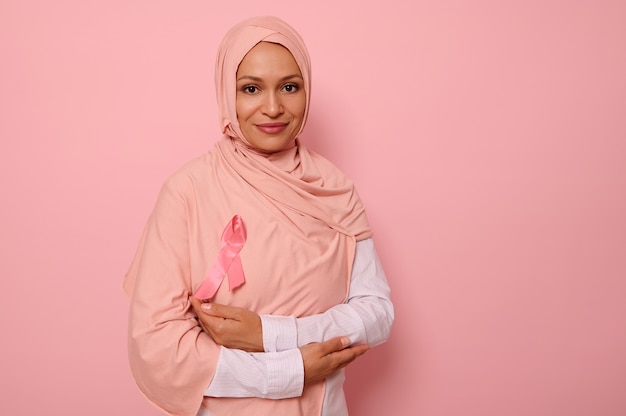 Splendida donna araba musulmana con la testa coperta in hijab, si erge su sfondo rosa con le braccia incrociate sul petto e indossa un nastro rosa, simbolo della giornata della consapevolezza del cancro al seno. concetto medico, copia spazio