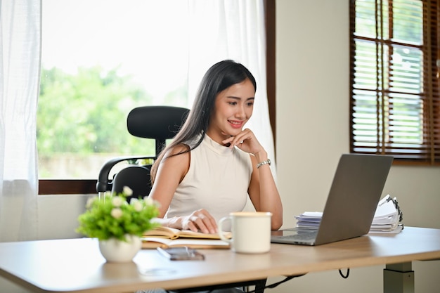 Великолепная азиатская деловая женщина сосредоточилась на своих задачах на ноутбуке, положив руку на подбородок