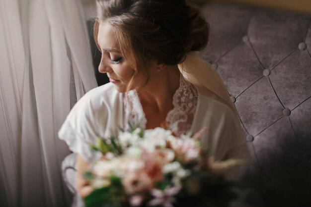 Photo gorgeous beautiful bride in silk robe holding modern bouquet and sitting on stylish gray sofa happy stylish bride with perfect makeup posing near window in soft light in the morning