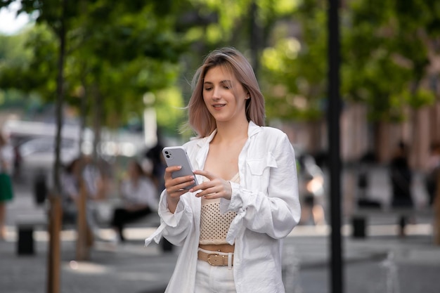 街の通りの背景でスマートフォンでブロンドの髪のメッセージを持つゴージャスな美しい若い女性は、街の通りでスマートフォンの会話をしているかわいい女の子