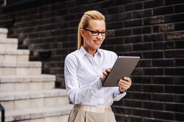 Gorgeous blond fashionable smiling businesswoman standing outdoors and using tablet