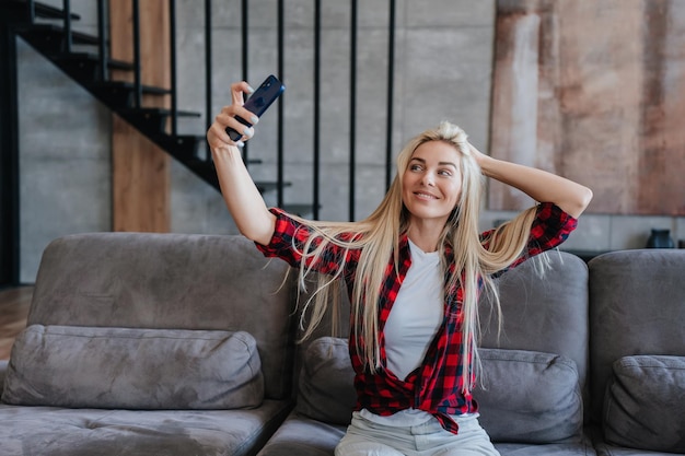Gorgeous blonde caucasian woman in plaid shirt holding phone makes selfie toothy smiling sitting on cozy sofa at home Beautiful model relaxing at weekend Cheerful Swedish girl using cellphone