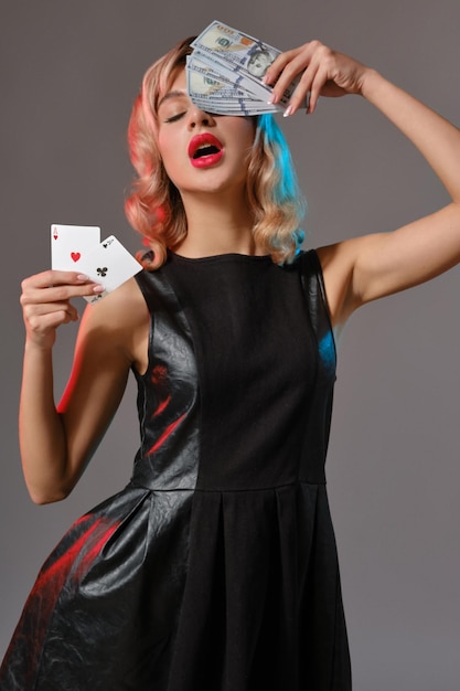 gorgeous blonde girl with bright makeup in black stylish dress is holding some money and cards while posing against gray studio background gambling entertainment poker casino closeup