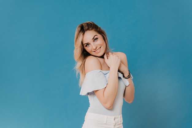 Gorgeous blonde wearing casual with folded hands on her chest, posing at studio, smiling broadly, looking with tenderness being in good mood, over blue background with copy space. Happy people concept