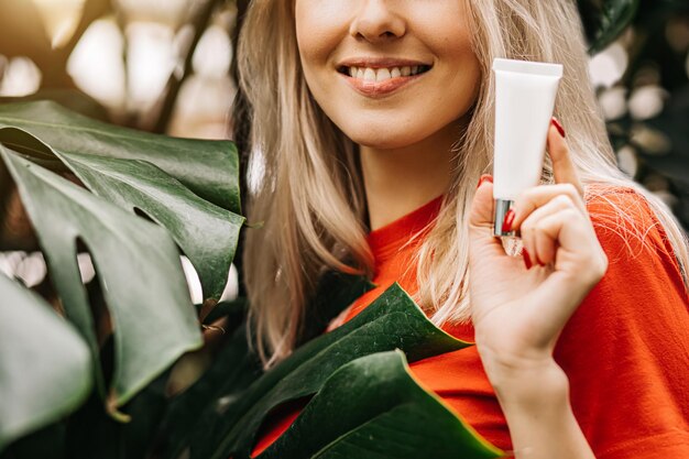 Gorgeous blonde woman holding tube of cosmetic product, eye
cream, sunscreen in the foreground in focus standing on tropical
plants background. beauty, cosmetics, advertising concept. cropped
photo