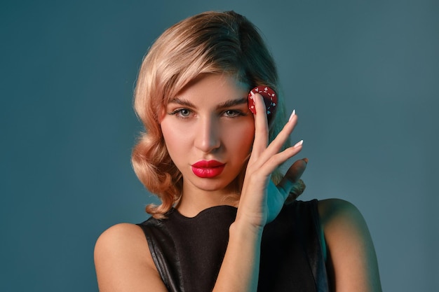 Foto splendida donna bionda con trucco luminoso in abito di pelle nera tiene in mano un chip rosso e posa su sfondo blu studio concetto di gioco d'azzardo intrattenimento poker casinò primo piano