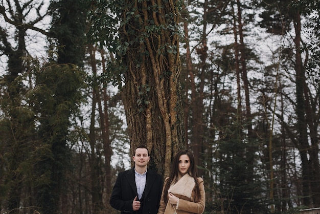 Gorgeous bride in coat and stylish groom posing under trees in\
winter forest happy wedding couple hugging in winter snowy park\
romantic sensual moment of newlyweds