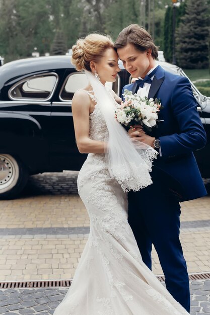 Gorgeous bride in vintage white wedding dress with bouquet hugging with handsome groom in blue stylish suit newlywed couple portrait outdoors