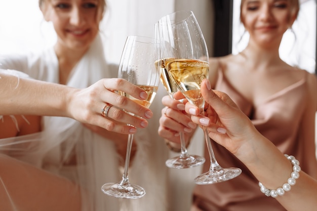 Gorgeous bride with best bridesmaids are holding glasses and drinking champagne in hotel bathroom near the window.