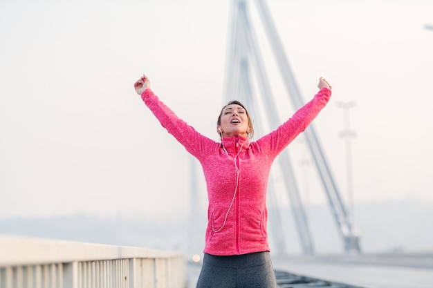 Gorgeous brunette lifting arms in the air and feeling free after running on the bridge. Winter time, healthy lifestyle concept.