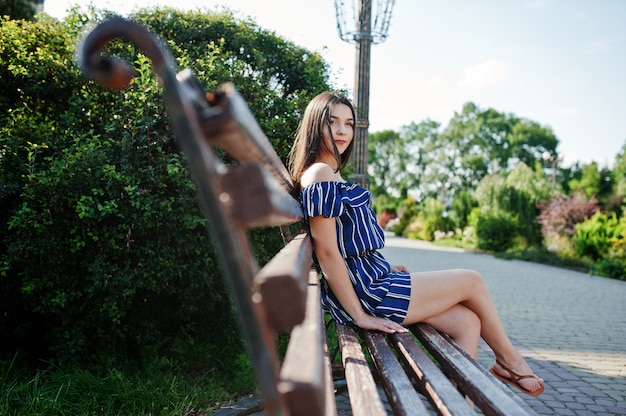 Gorgeous brunette woman sitting on bench at street of city wear on blue striped dress.