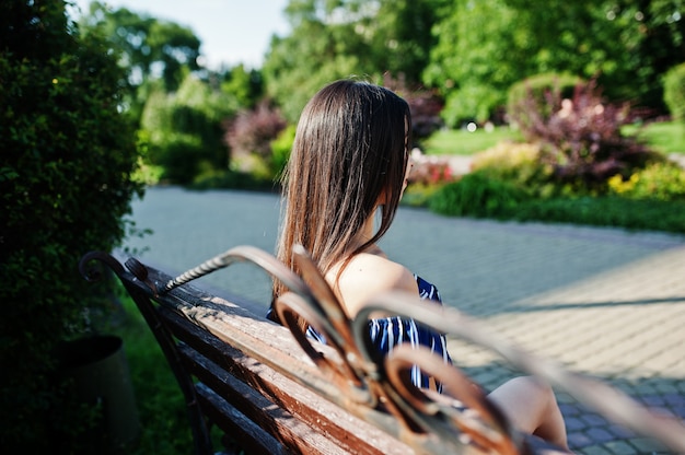 Gorgeous brunette woman sitting on bench at street of city wear on blue striped dress.